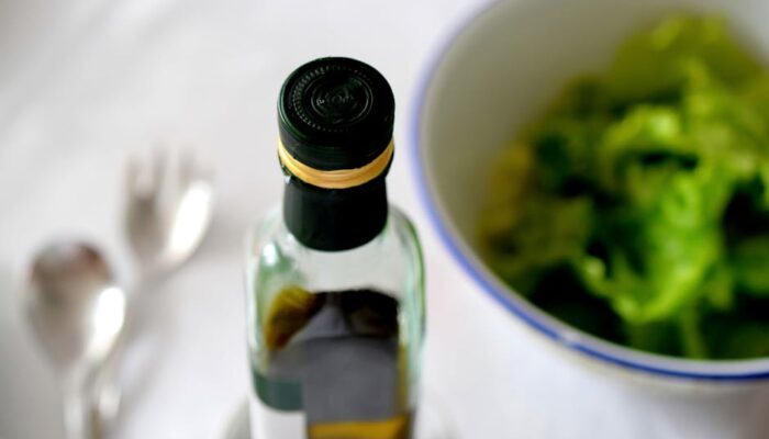 A high-angle view of an olive oil bottle alongside a fresh green salad and cutlery. - Photo by Marco Antonio Victorino on Pexels