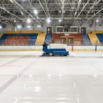 A Zamboni ice resurfacer operating in an empty indoor ice skating rink with colorful seating. - Photo by Pavel Danilyuk on Pexels