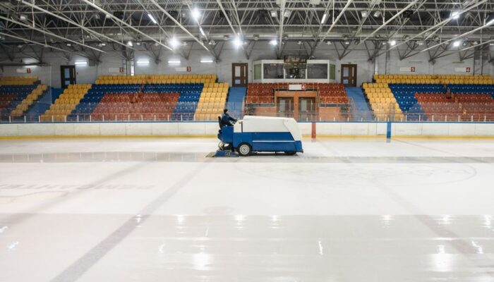 A Zamboni ice resurfacer operating in an empty indoor ice skating rink with colorful seating. - Photo by Pavel Danilyuk on Pexels